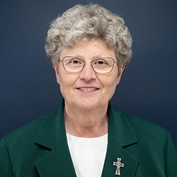 Sister Laura Reicks smiles into the camera against a dark blue background.